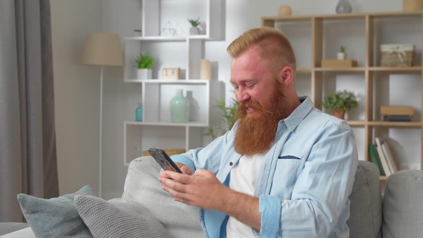 Addicted to technology young handsome redhead man resting on cozy sofa, involved in using smartphone software application, web surfing information, communicating in social networks alone indoors. | Shutterstock HD Video #1099353531