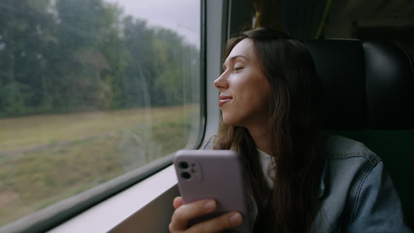 A smiling young woman enjoys beautiful views from the window while riding on the train. The brunette holds a mobile phone in her hands Royalty-Free Stock Footage #1099354155