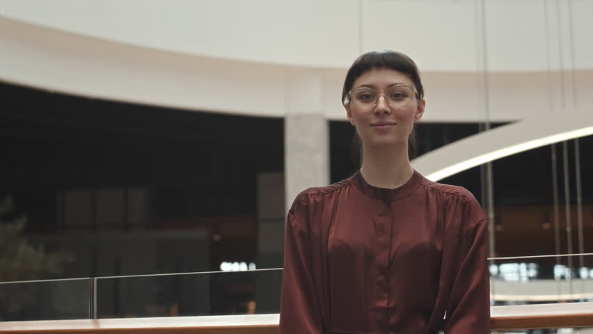 Medium portrait of young confident Caucasian woman in eyeglasses posing with hands folded and smiling at camera standing at contemporary business centre