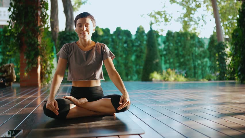 Sunny short-haired woman meditating with closed eyes in the lotus position outdoors Royalty-Free Stock Footage #1099356525