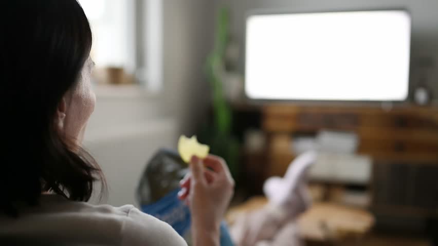 Woman watching television at home and eating chips | Shutterstock HD Video #1099356589