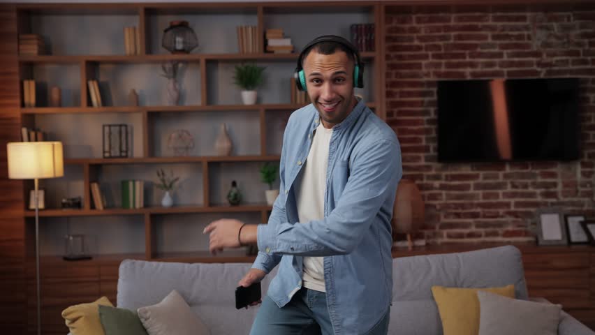 African American man in casual clothes dancing at home living room near couch and holds smartphone in his hands wears headphones listening to music online on radio dancing actively moving having fun