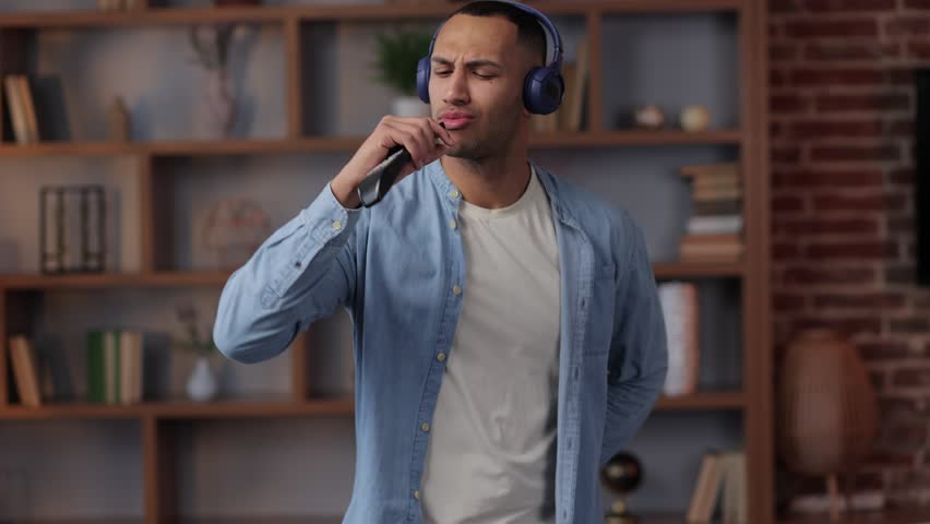 Portrait of attractive man in headphones dancing and singing into a remote control instead of a microphone at home in slow motion. Positive guy gesturing to music on modern room. | Shutterstock HD Video #1099357239