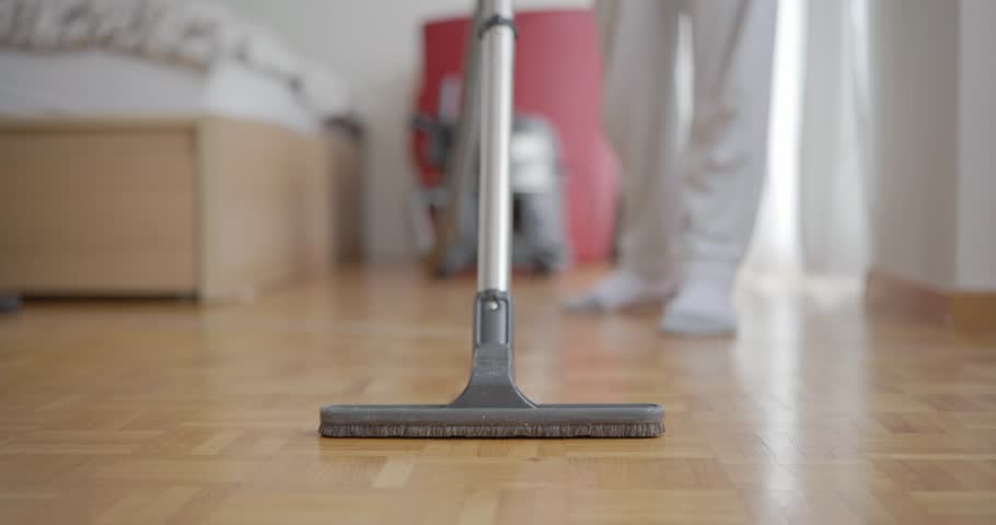 Unrecognizable blurry out of focus male, using a vacuum cleaner brush and extension rod on laminated wooden tile surface inside apartment. Close, upwards looking shot, shallow depth of field. | Shutterstock HD Video #1099357719