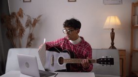Young man playing acoustic guitar and watching online lesson on laptop while practicing at home. Online training, online classes concept