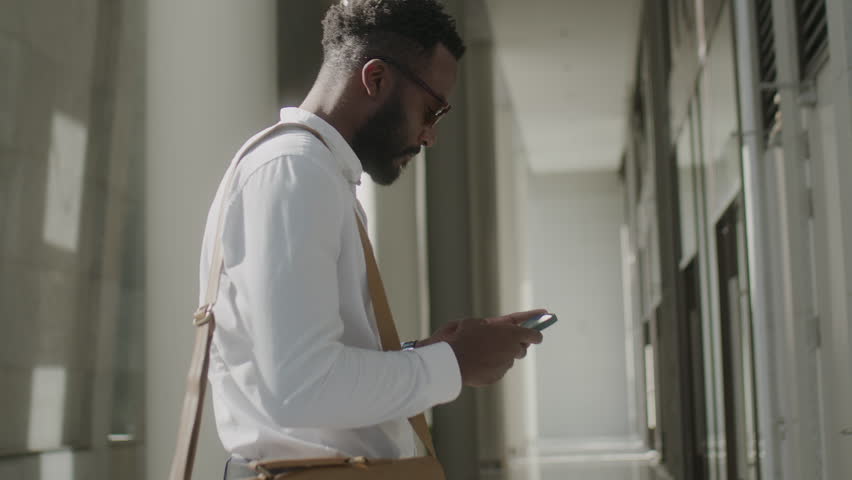 African American man in white shirt with bag and glasses standing in a open space hall and surfing the internet on mobile phone, sunlight  | Shutterstock HD Video #1099361907