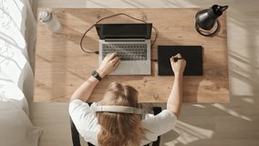 Top down view of girl at desktop with laptop and graphics tablet. Young blonde girl draws sketches or illustrations, put on headphones and drink water from her bottle. Freelance work during day room. - Powered by Shutterstock - Get 15% off with code: PIKWIZARD15