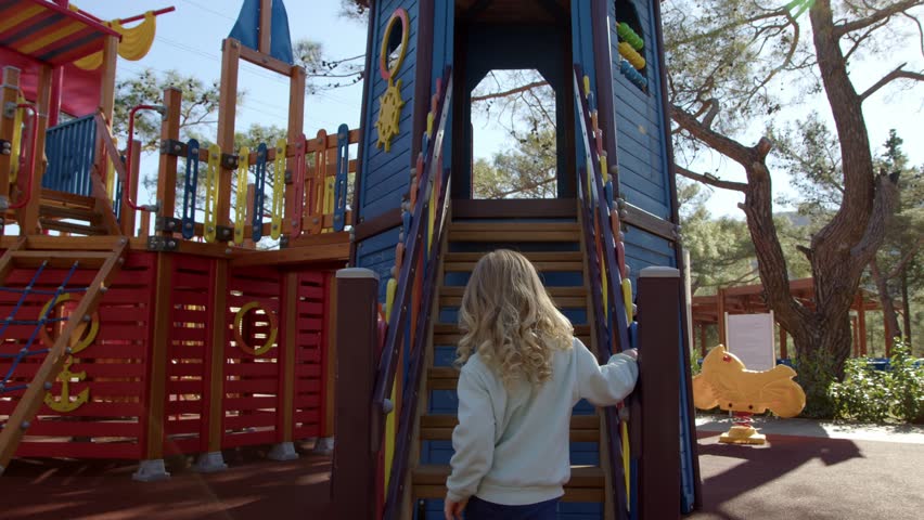 Blonde curly-haired girl climbs up stairs to colored playground. happy child plays alone and enjoys game. girl climbs up to slide down children slide. | Shutterstock HD Video #1099368695