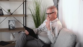 Family video call. Virtual chat. Online communication. Relaxed cheerful middle-aged man in glasses waving hi on tablet camera sitting on couch in light home living room interior.
