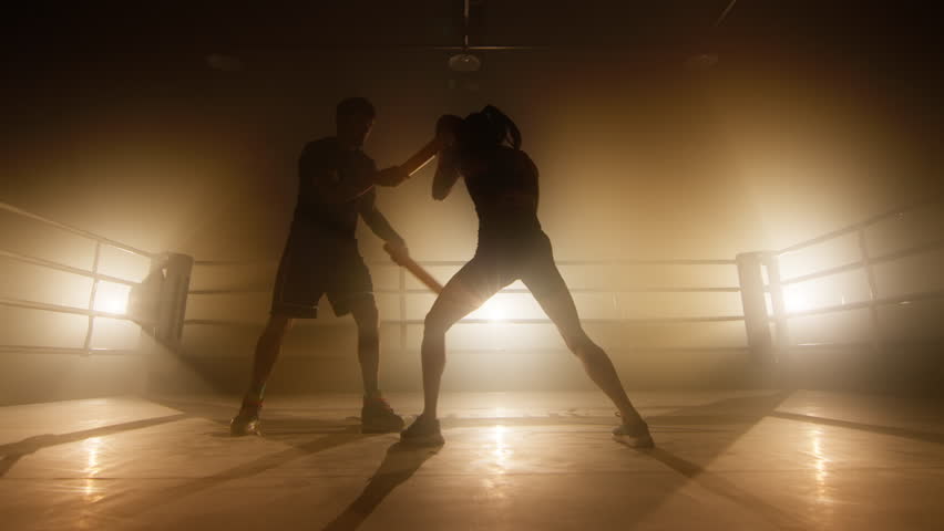 Silhouettes of man and woman having intensive workout indoors. Close-up view of female kickboxer training with her coach at boxing club. High quality 4k footage in golden orange yellow foggy light | Shutterstock HD Video #1099370395