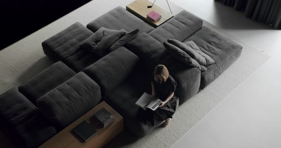 A woman is sitting on the couch and reading a book. Luxury Modern House Interior With Corner Sofa and Bookshelf. Fashionable furniture. Minimalist Home Interior. Cozy Modern Furniture Design. Royalty-Free Stock Footage #1099371651