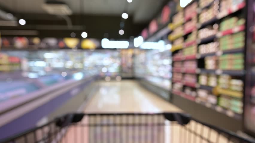 Blurred and trolley shopping cart moving through food product shelves interior defocused background in the supermarket. person pushing a shopping cart in a supermarket store in slow motion shot.