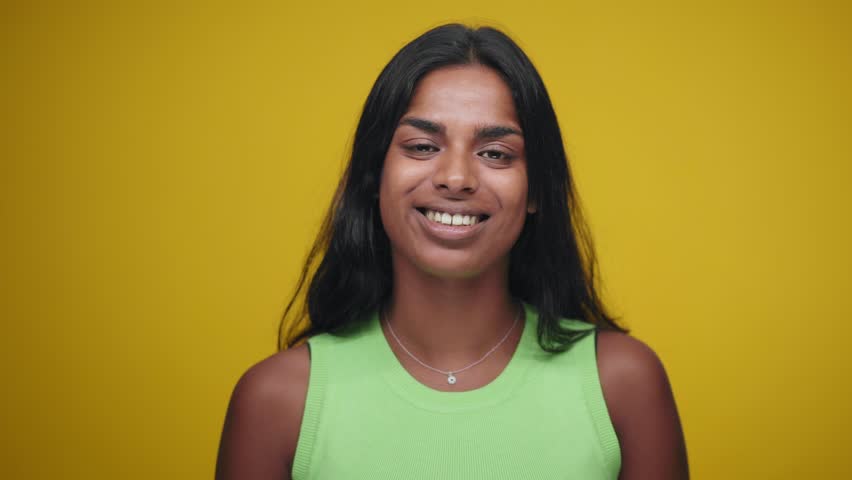 Portrait of Young Indian Woman Looking at Camera and Smiling in Color Studio Shot. Happy adult girl isolated on yellow background having fun Closeup. Indian person with beautiful skin tilts head. | Shutterstock HD Video #1099373061