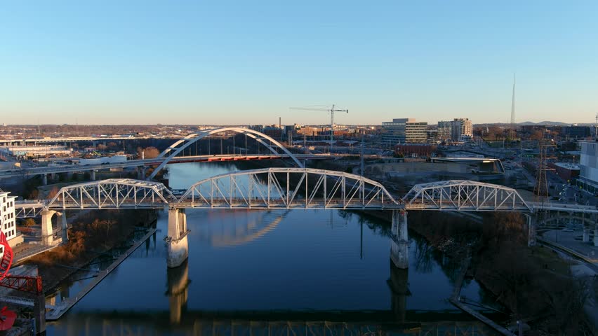 Nashville Tennessee USA - 1 16 2023: aerial footage of the rippling green waters of the Cumberland River and the John Seigenthaler Pedestrian Bridge with winter trees and blue sky Royalty-Free Stock Footage #1099376001