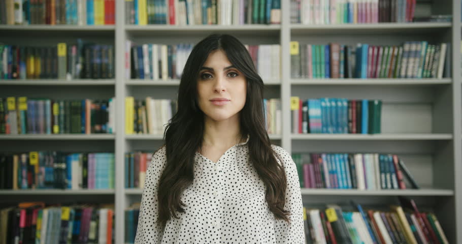 Arab young woman tells about spending day in library and demonstrates sincere emotions. Lady stands against racks with books in library