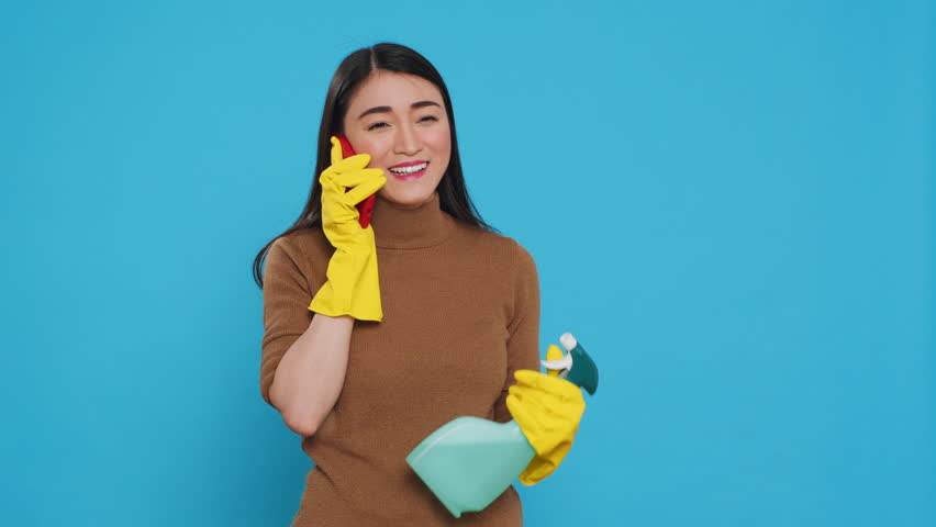 Cheerful housewife wearing protective gloves while holding chemical spray bottle talking at smartphone with remote friend, standing in studio over blue background. Housekeeping and cleaning concept.