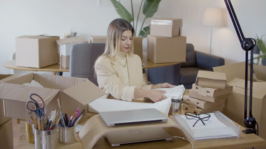Young Caucasian woman wrapping up piece of clothing, getting it ready for delivery to customer and packing item in cardboard parcel. Happy young businesswoman at work. Startup, online store concept | Shutterstock HD Video #1099387965