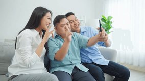 Asian attractive parents and young son using phone video call at home. Happy family, Beautiful loving mother and father sitting on sofa with young man then talk virtual online on smartphone in house.