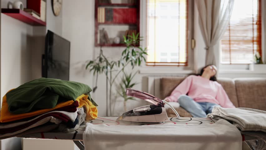 Close up of stack of clothes and steaming hot iron left on ironing board. In backgroung defocused housewife sleeps on sofa. Home chores. | Shutterstock HD Video #1099390913