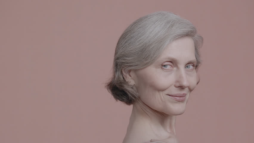 Portrait of beautiful 60s grey-haired mature senior adult female posing without makeup against light brown background. Studio shot, soft lighting. No make up, clean skin. Shot with ARRI Alexa Mini LF | Shutterstock HD Video #1099391487