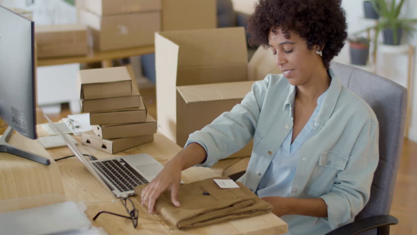 Serious Black woman checking clothing paper tag and barcode on computer, and then packing item in cardboard parcel. Female webstore owner making delivery preparations. Startup, online store concept | Shutterstock HD Video #1099392265