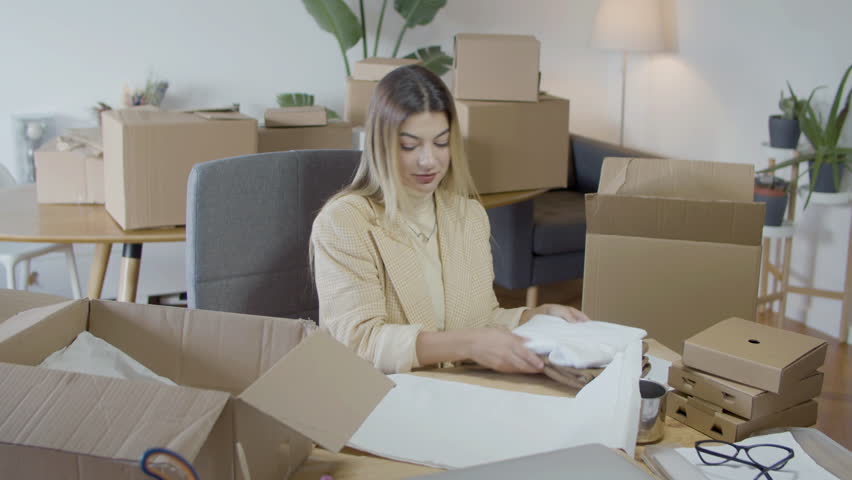 Elegant Caucasian woman packing purchased clothing, putting product in cardboard parcel. Successful young businesswoman drinking coffee and making notes. Startup, small business concept | Shutterstock HD Video #1099392273