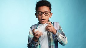 Cute little school boy with glasses playing in online video games on smartphone in horizontal landscape mode on isolated blue background Adorable kid with backpack having fun after lessons alone