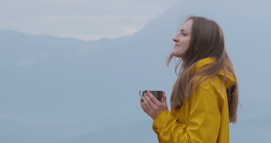 Beautiful smiling girl on top of mountain corrects fluttering hair from wind. Woman drinks hot drink on mountain background