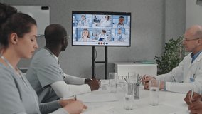 Group of modern medical workers sitting at table discussing treatment problems with colleagues on video call