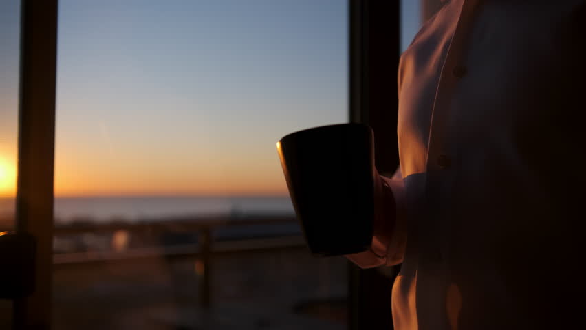 A man with a cup of hot tea looks out the window where the sun has risen. 