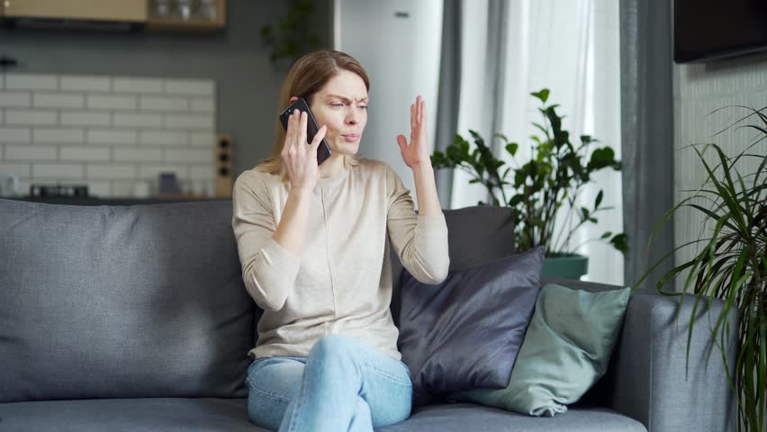 Irritated arguing blond woman yelling at cellphone while sitting on the couch at home Angry nervous young female talking on a smartphone expressing negative emotion Bad conflict conversation | Shutterstock HD Video #1099411687