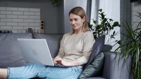 Beautiful woman working typing on laptop while sitting on sofa in living room at home. A female freelancer or student listens to an online conference in the kitchen, holding the device on her lap