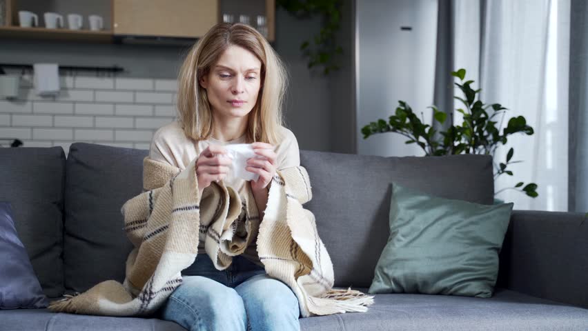Sick blond woman with cup of hot tea sitting on the couch under the warm blanket and has severe headache temperature at home Unhealthy female getting flu virus symptom Cold and fever concept | Shutterstock HD Video #1099411733