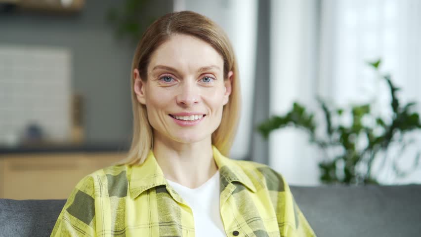 Close-up of caucasian adult woman looking at camera and smiling while sitting on sofa in living room. Happy lady with beautiful face posing for portrait indoors She turns her head with joyful emotions | Shutterstock HD Video #1099411745