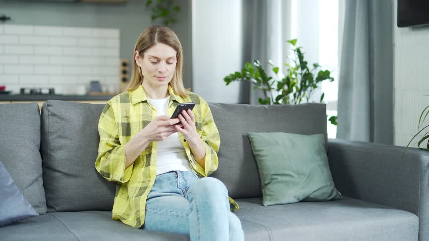 A young smiling girl is using a smartphone while sitting on the sofa. relaxing while chatting on mobile phone. Surfing social networks, checking news. a female is streaming a video on her mobile phone | Shutterstock HD Video #1099411761