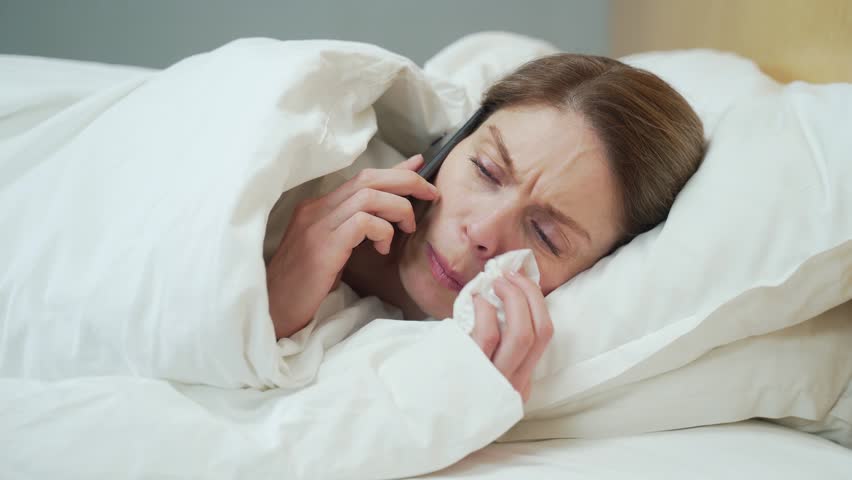 Sick blond woman blowing running nose sneezing and coughing laying on the warm bed in the bedroom Unhealthy female under the blanket getting flu virus symptom Cold and fever concept | Shutterstock HD Video #1099411767
