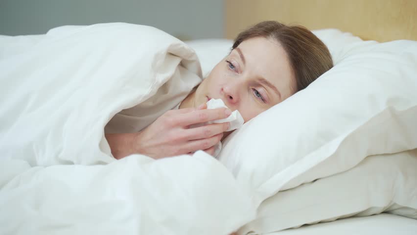 Sick young blond woman blowing running nose sneezing and coughing laying on the warm bed in the bedroom Unhealthy female under the blanket getting flu virus symptom Cold and fever concept | Shutterstock HD Video #1099411771