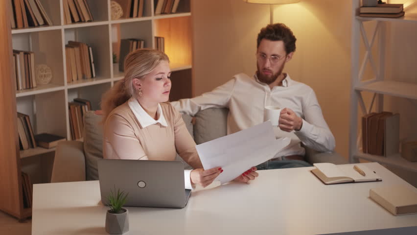 Bad work. Displeased woman. Colleagues quarrel. Angry female manager showing wrong documents to relaxed careless man sitting sofa drinking coffee in light room interior. | Shutterstock HD Video #1099414611