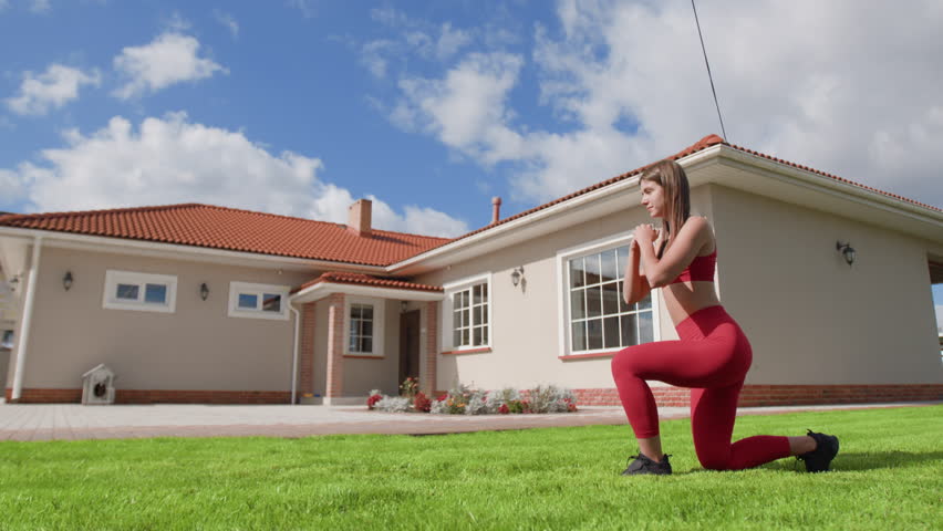 Woman doing lunges in red tracksuit outdoors. Caucasian female athlete training her hips on sunny morning within beautiful nature. High quality 4k footage