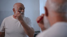 Reflection in mirror of senior Caucasian man shaving stubble on cheek with razor. Concentrated confident old retiree styling white beard at home in bathroom. Hygiene and lifestyle concept - Powered by Shutterstock - Get 15% off with code: PIKWIZARD15