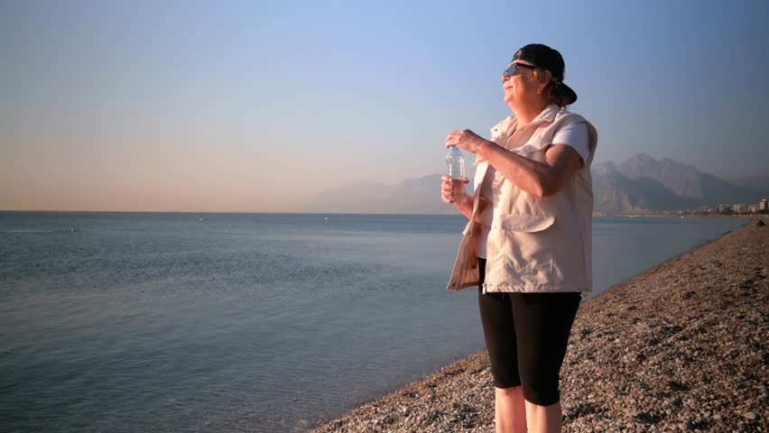 Woman in cap and sunglasses drinking water from bottle on blue sky background. Tourist tired. Outdoors