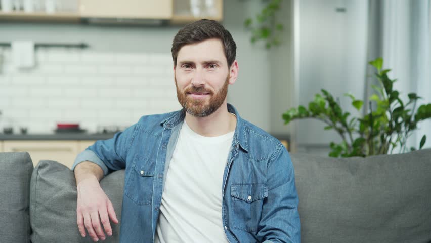 Close-up portrait of mature сaucasian man with beard smiling and looking at camera while sitting on couch in living room Confident handsome male in casual clothes relaxing on sofa in kitchen at home | Shutterstock HD Video #1099430633