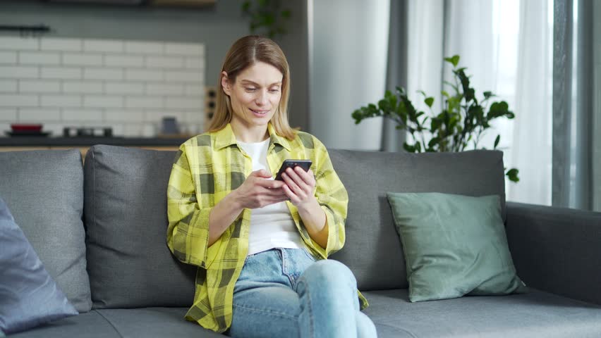 Caucasian woman using smartphone while sitting on sofa in living room at home. Concentrated middle-aged female checking email, texting or browsing social media while holding device indoors | Shutterstock HD Video #1099430637