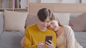 Asian young couple lover sit on couch or sofa looking short video clips in social media having fun and laughing together. Happiness family spending time with good moment together in holiday at home.