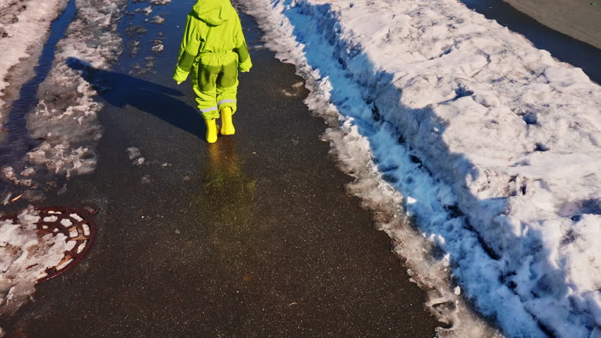 Back view of unrecognizable child in yellow waterproof rubber boots walking on thawing snow in wet sidewalk next to blackened icy snowdrifts
