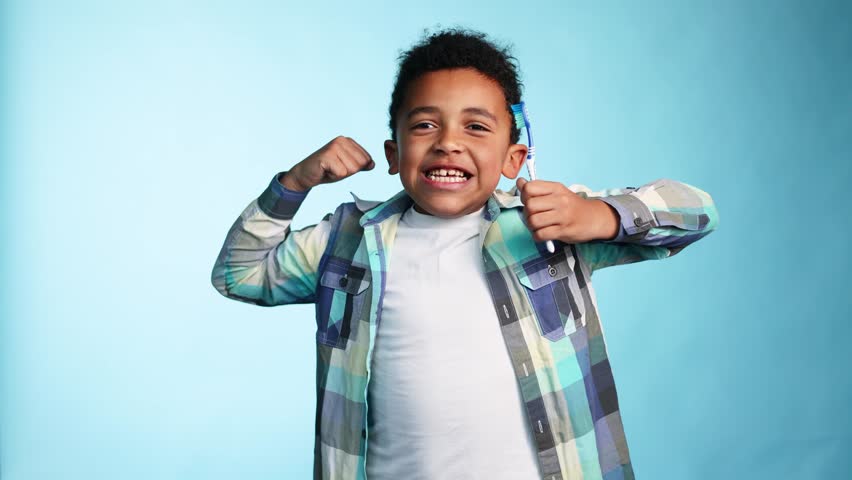 Cute little african american boy with teeth brush in his hand showing his biceps power to the camera on isolated blue background Adorable kid motivate other brushing teeth Daily healthcare concept | Shutterstock HD Video #1099440367