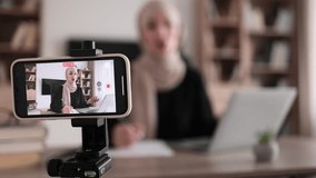Focus on screen of phone of cheerful young muslim woman in hijab recording creative video blog for her social networks, media for followers while working with laptop and writing report.