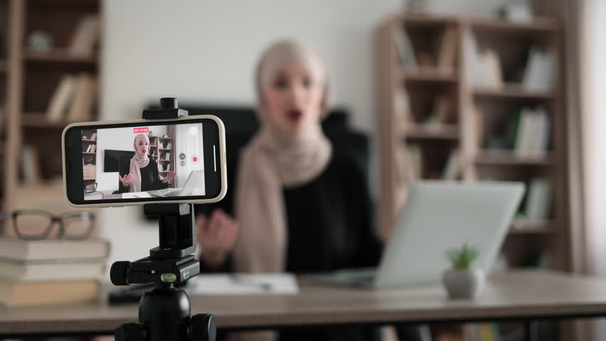 Focus on screen of phone recording video blog of beautiful muslim woman in hijab talking while looking at modern smartphone. Female blogger sitting at home and doing live stream. | Shutterstock HD Video #1099445121