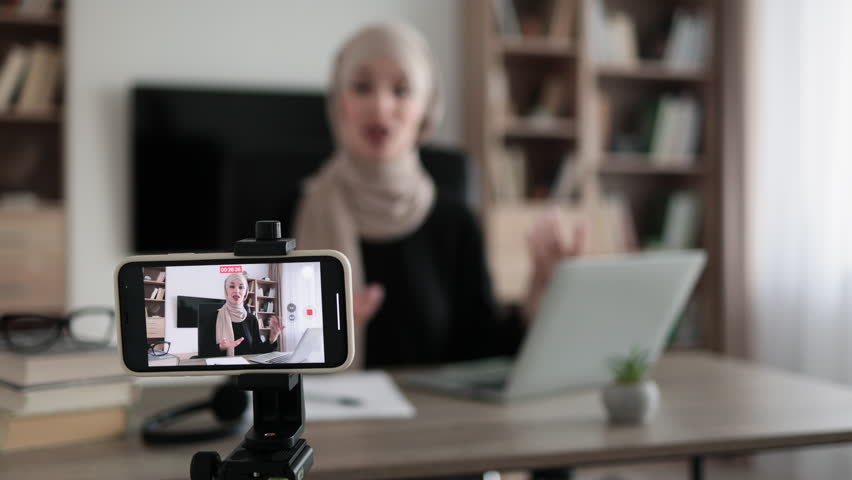 Focus on screen of phone of happy arab woman sitting at desk with books, notes and laptop talking with followers. Female tutor working at home during distance learning and showing sign ok. | Shutterstock HD Video #1099445139