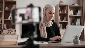 Confident muslim female in headscarf sitting at desk working on laptop and recording video on digital smartphone. Concept of freelance and modern technology.
